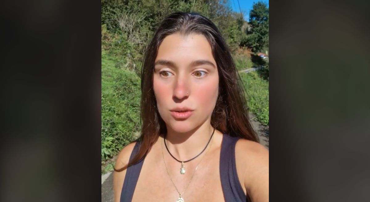 Una chica con camiseta de tirantes negros dos collares a distinto nivel y pelo castaño y largo paseando por un camino rodeado de prados