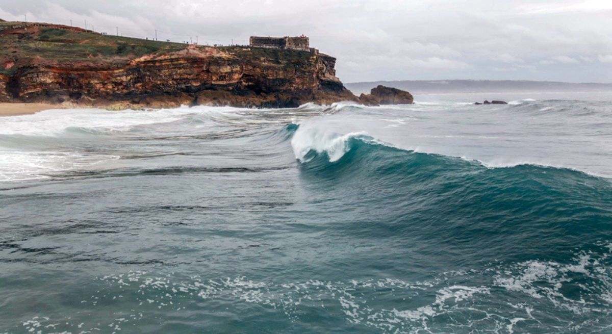 Playa con oleaje. Se puede ver un peñón al fondo y una ola rompiendo en el mar