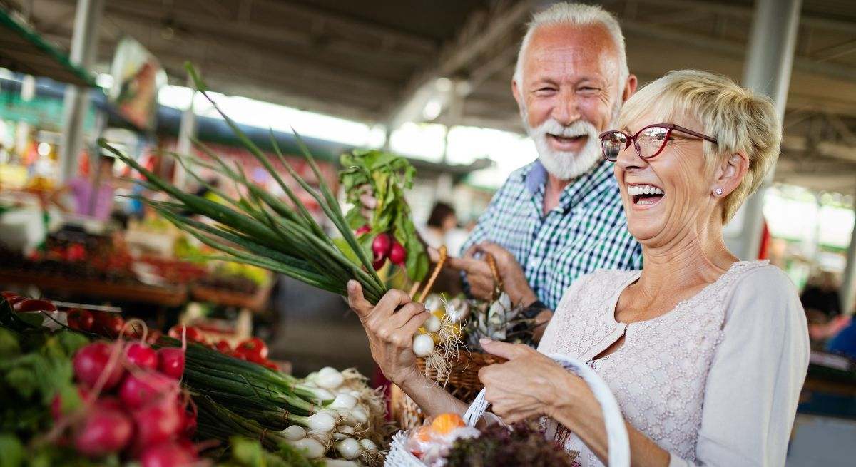Una pareja de señores mayores comprando frutas y verduras en un mercado y riéndose mucho
