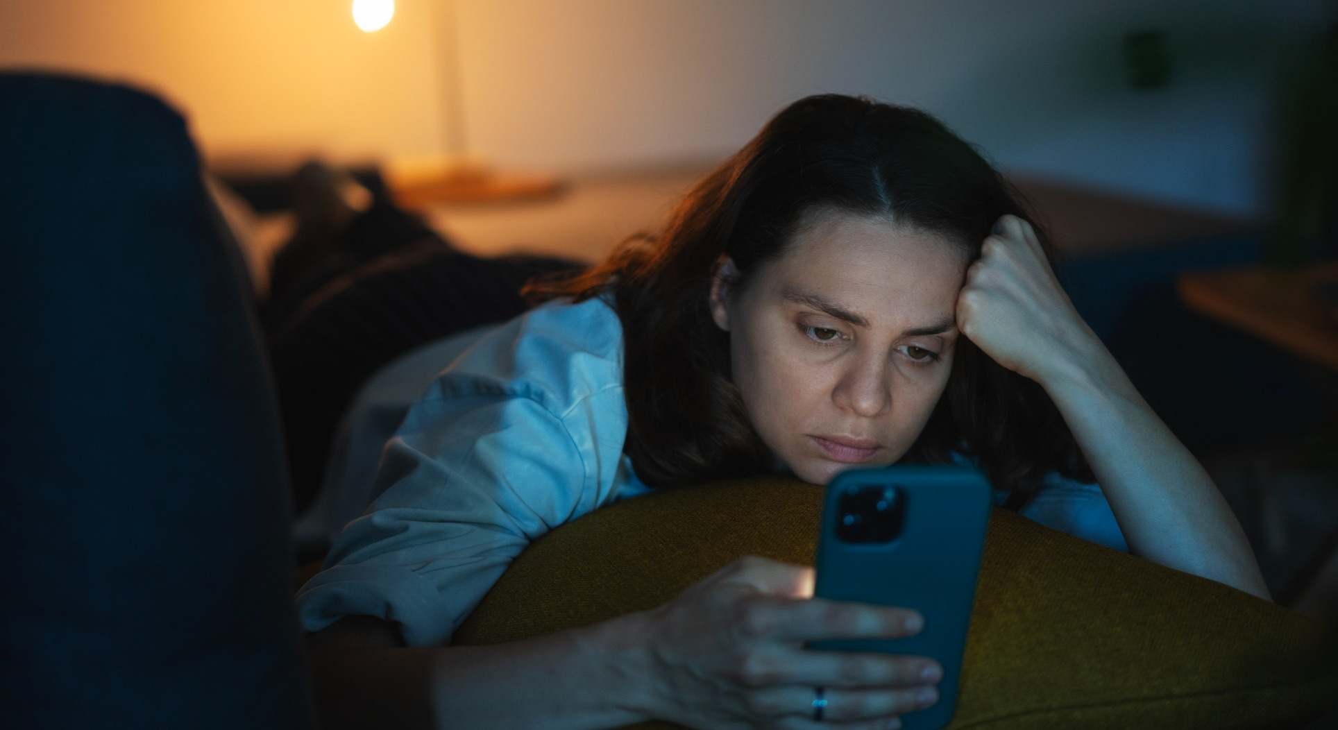 Mujer pensativa mirando el móvil sobre la cama con luz tenue