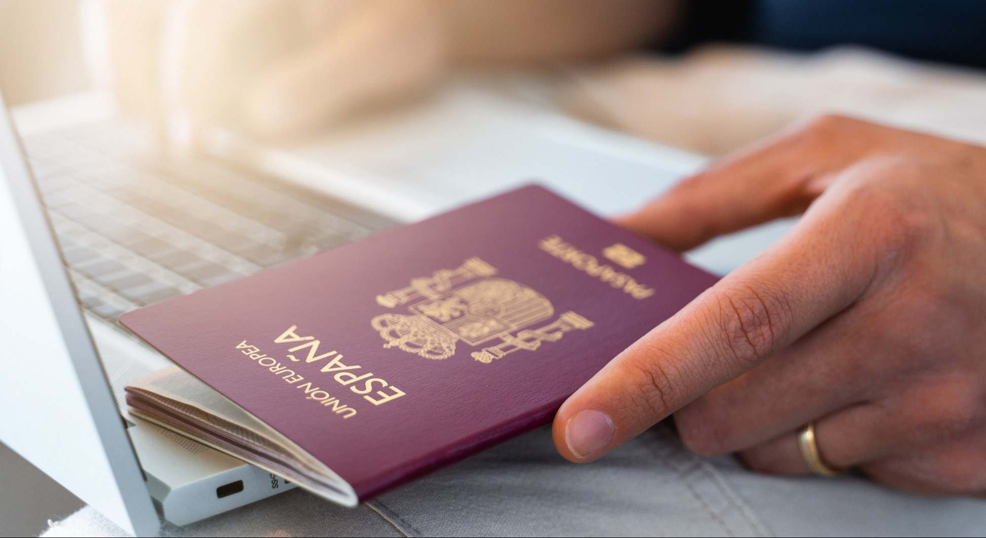 Primer plano de mano masculina sosteniendo un pasaporte español mientras se utiliza el ordenador portátil