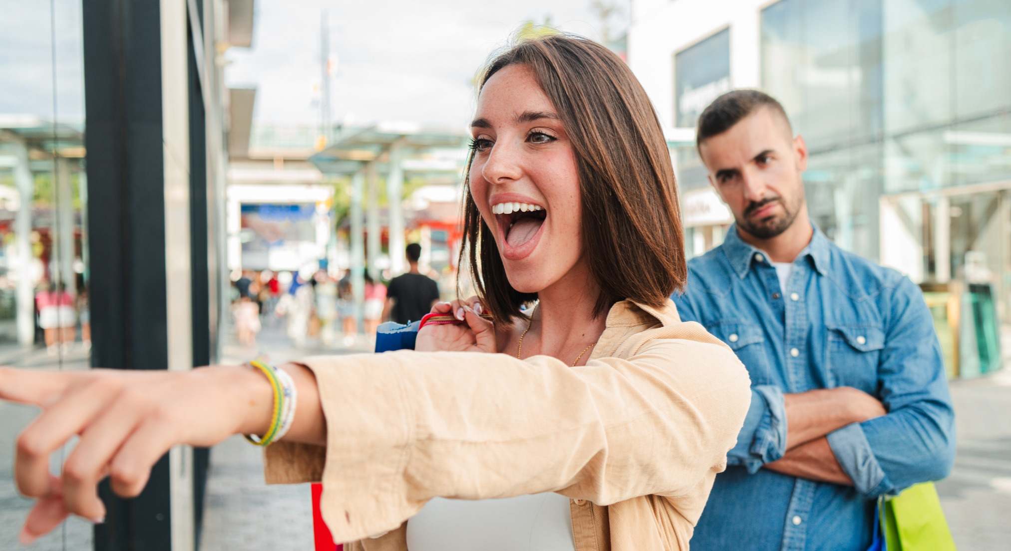 Mujer sonriente señalando un escaparate con su pareja detrás disgustada