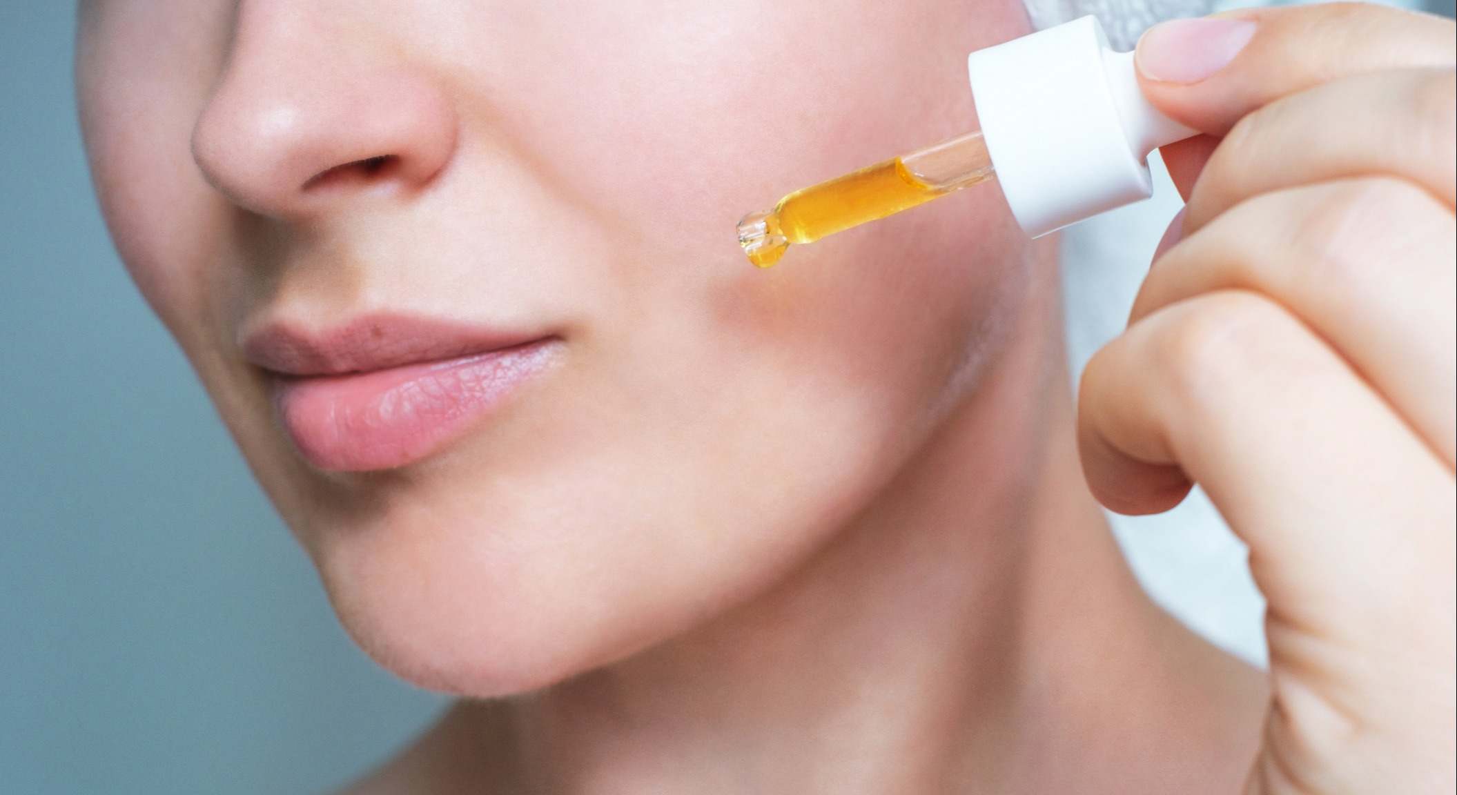 Rostro femenino y gotero en primer plano de suero hidratante sobre fondo gris.