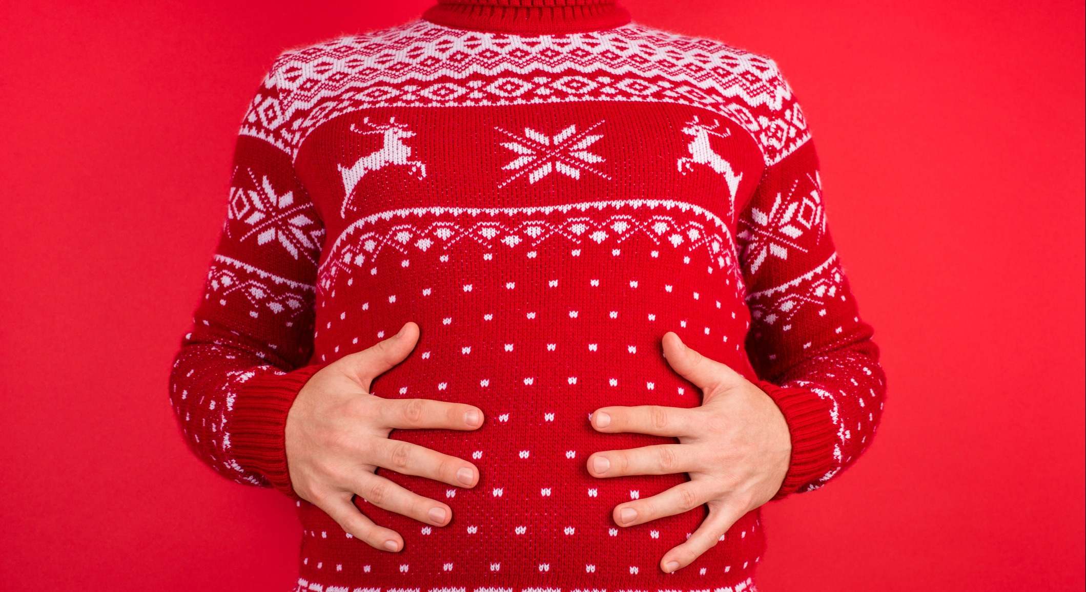 Foto de un hombre con suéter rojo de invierno con ciervos sosteniendo su gran barriga aislada en el fondo rojo