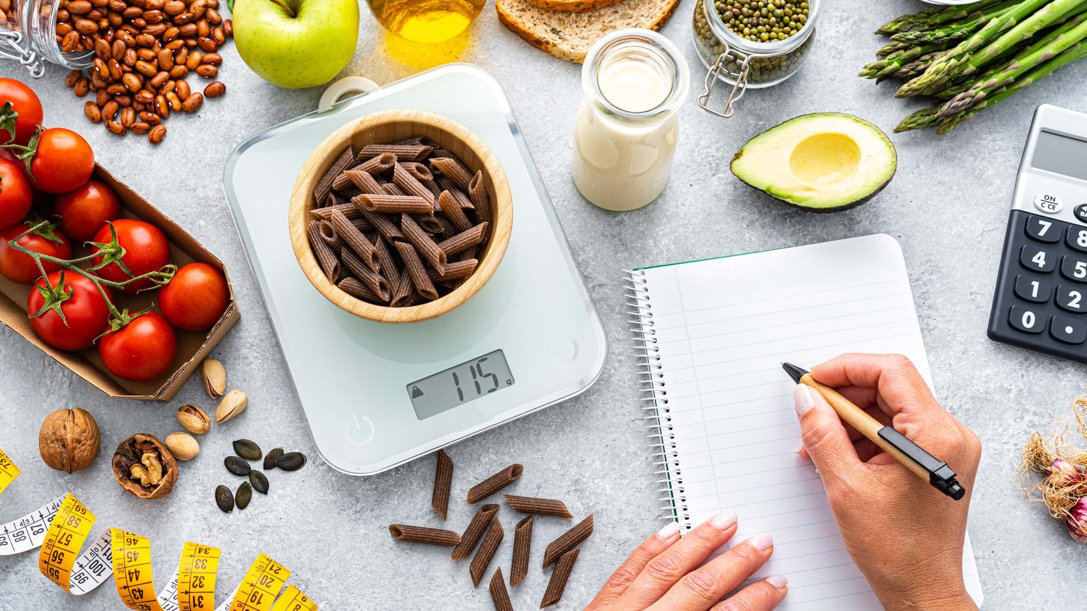 Mesa con alimentos saludables, una báscula, una persona anotando en una libreta y una calculadora