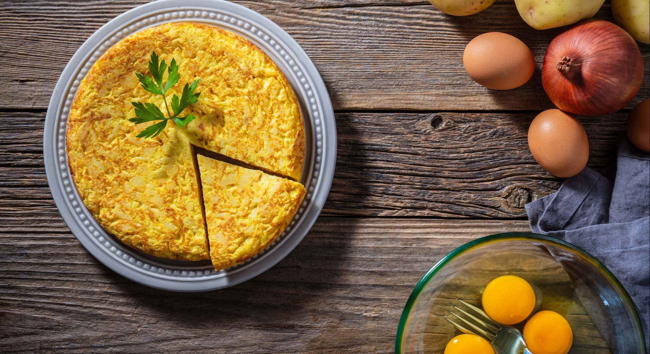 Una tortilla de patatas en una mesa de madera con un bol de huevos y cebolla