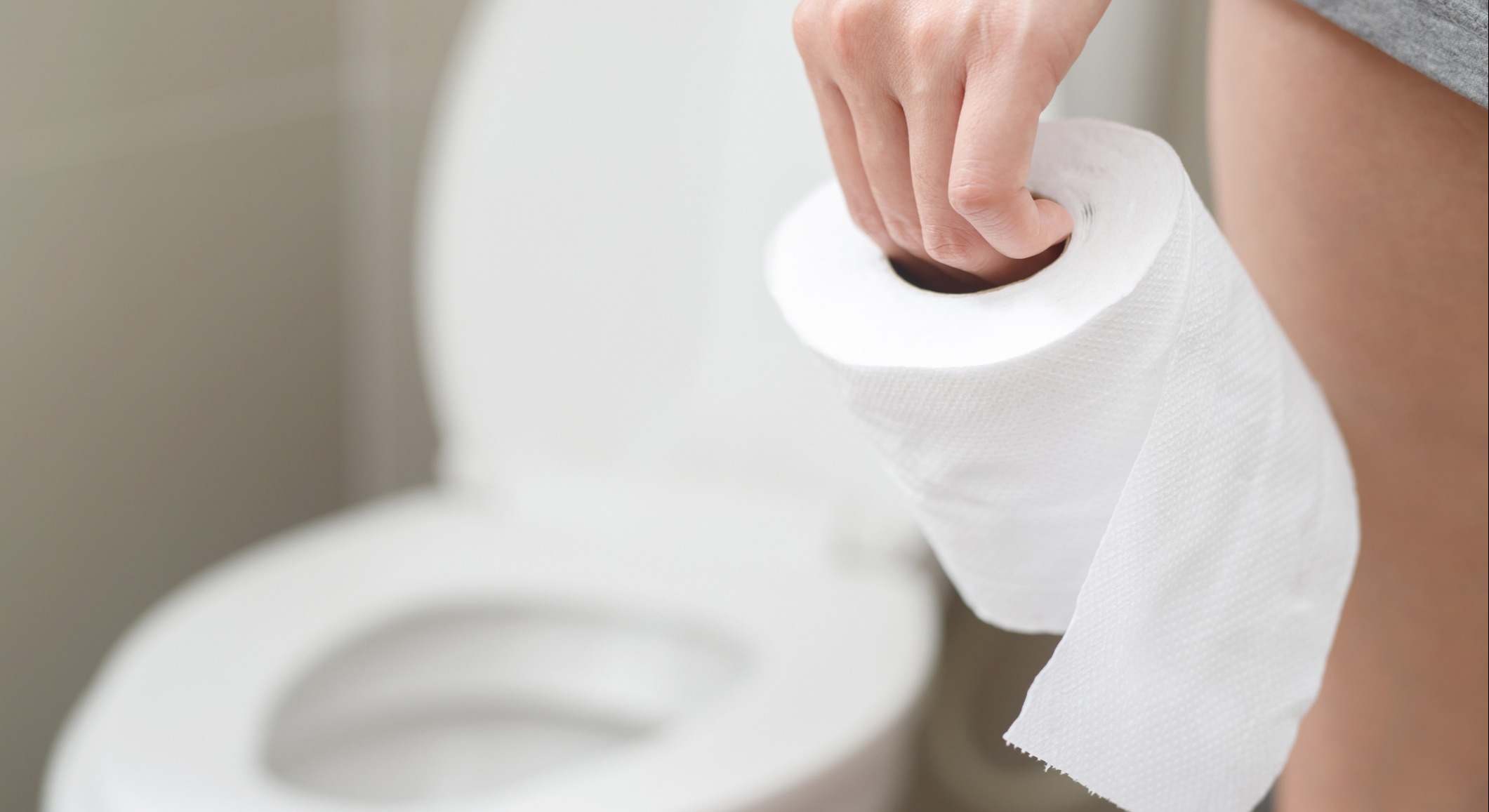 Persona sujetando con la mano un rollo de papel higiénico con un váter de fondo