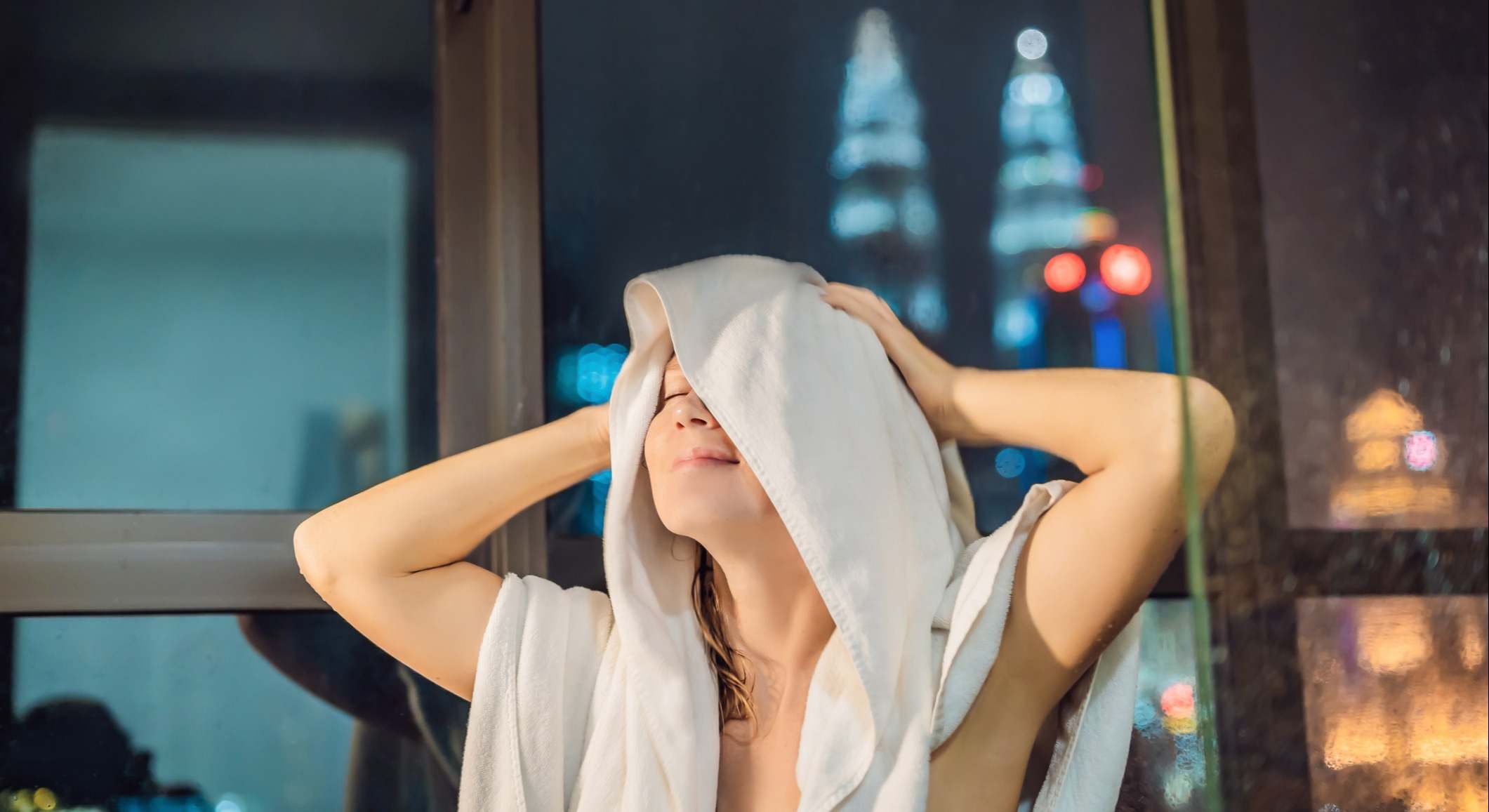 Mujer se seca el pelo después de una ducha con un fondo de una ventana con una vista panorámica de la ciudad nocturna