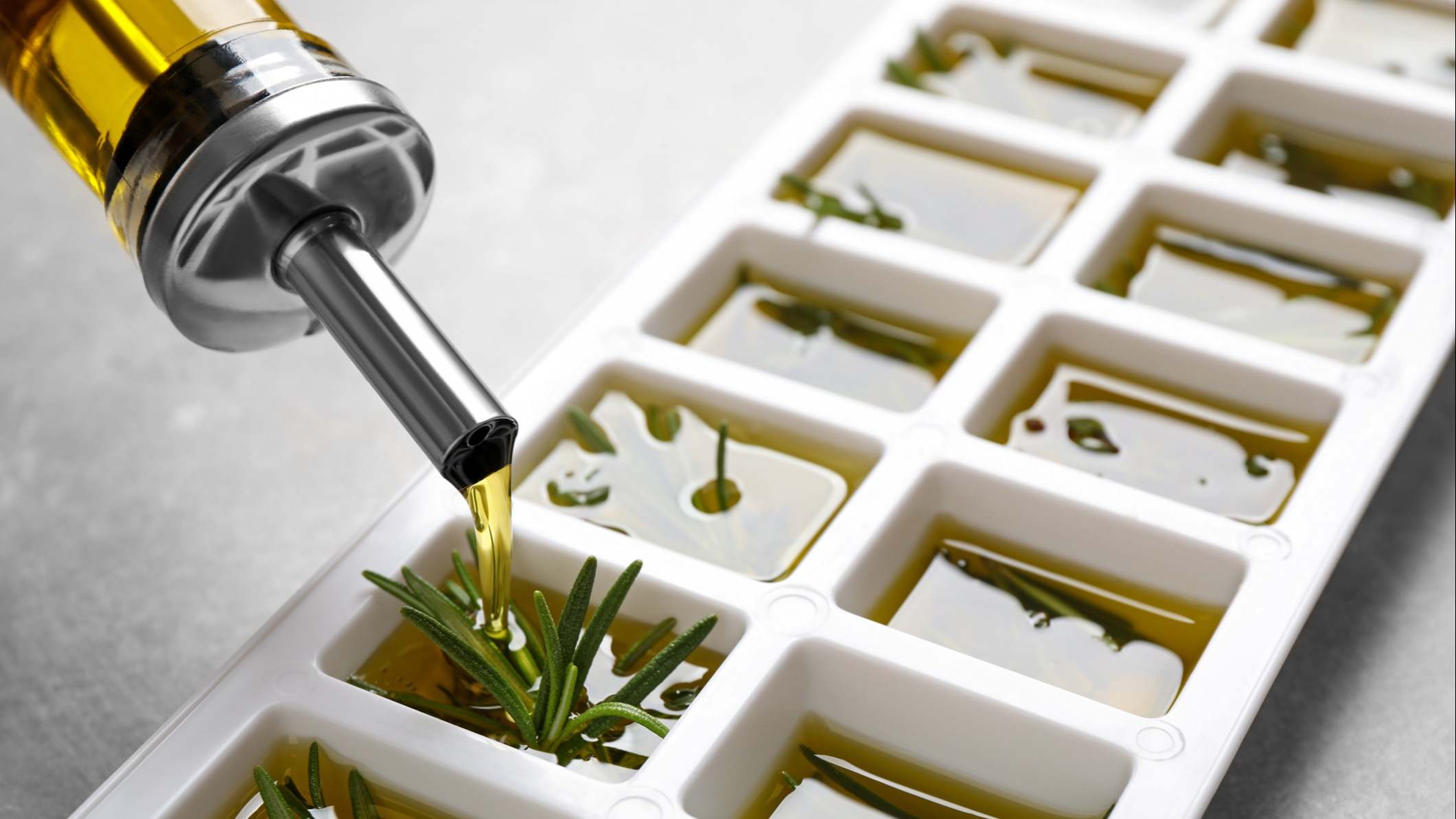 Aceite de oliva vertido en bandeja de cubitos de hielo con romero fresco en la mesa gris