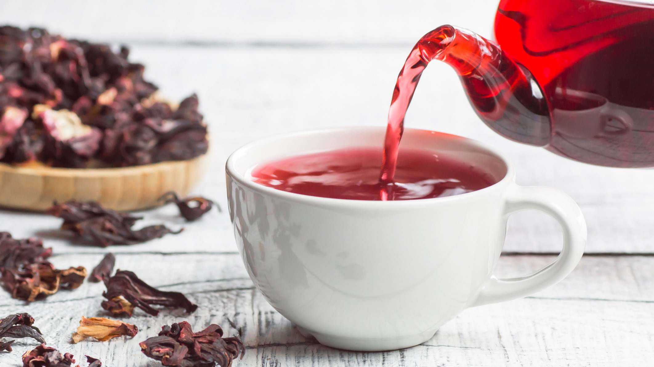 Taza blanca de té de hibisco que se vierte de la tetera con flores de hibisco secas sobre fondo de madera blanca