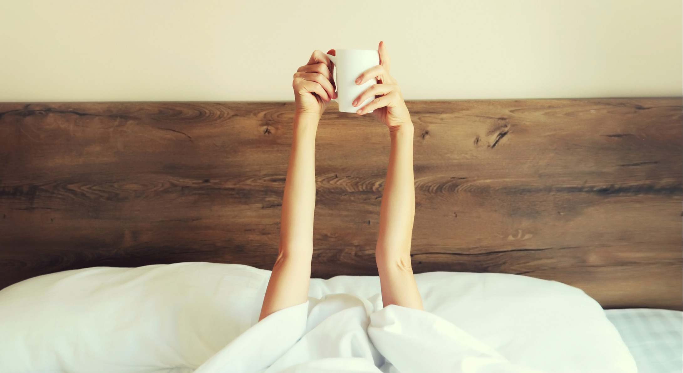 Mujer con los brazos en alto sujetando una taza dentro de la cama