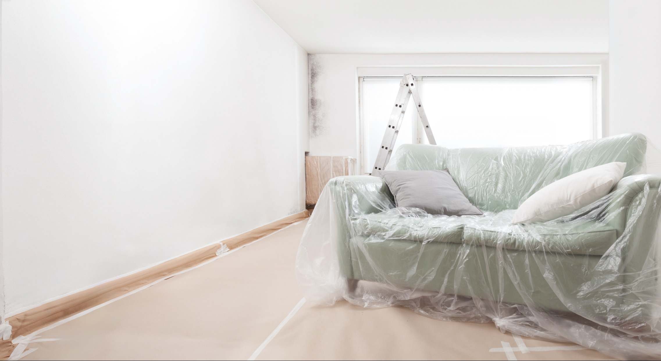 Habitación vacía en renovación con pared blanca, escalera y sofá cubierto con láminas de plástico de pintor frente a una ventana luminosa