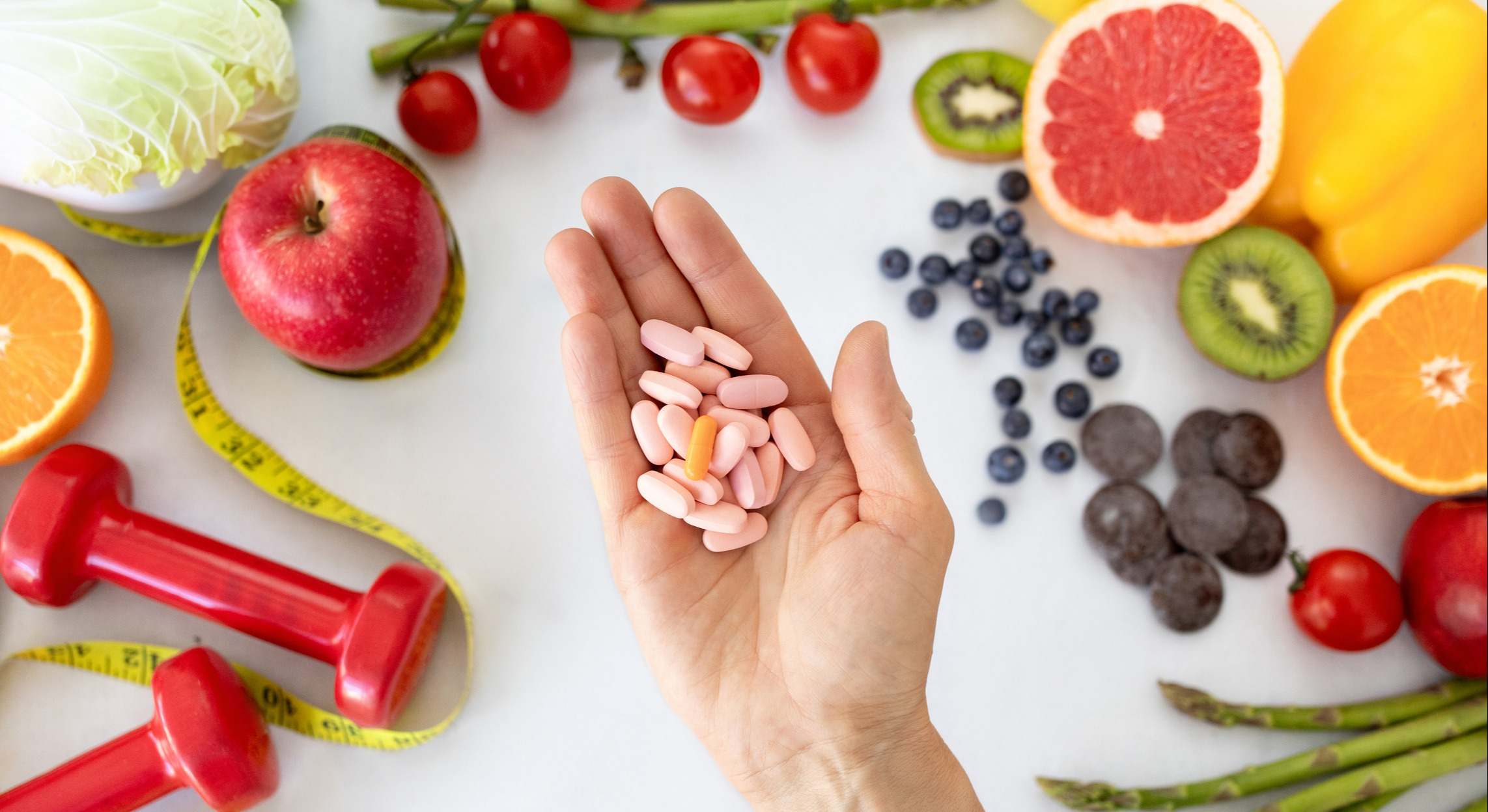 Mano sosteniendo píldoras, con pesas, frutas y una cinta métrica de fondo