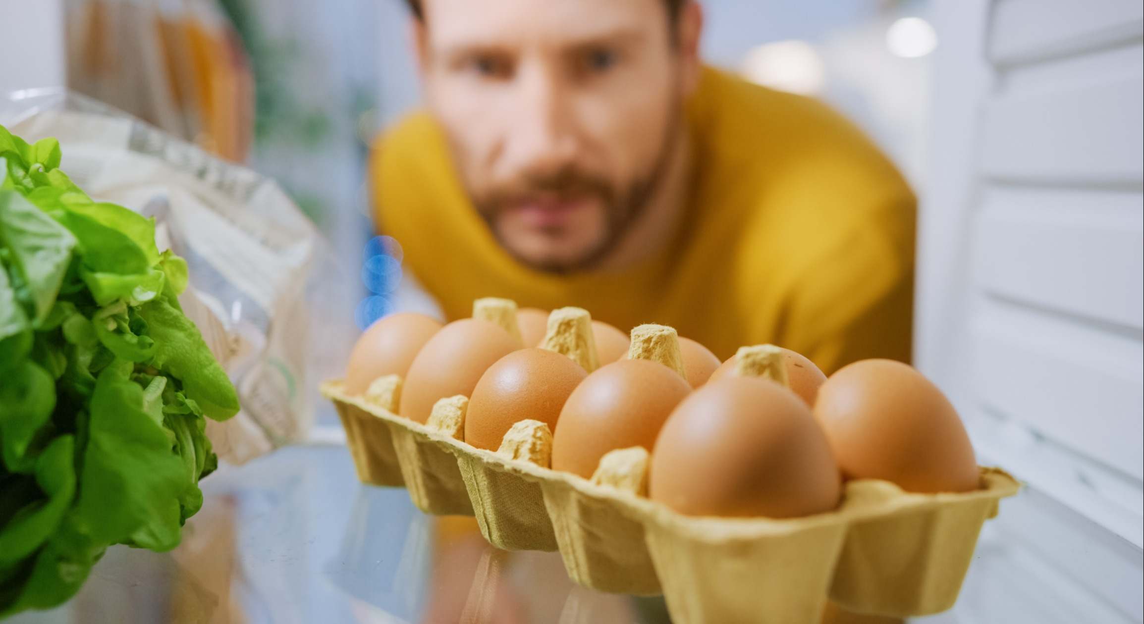 Disparo desde el interior de la nevera de la cocina: Handsome Man Abre la puerta de la nevera, mira dentro y piensa en tomar los huevos de la caja de los huevos. Point of View POV Shot from Refrigerator lleno de comida saludable