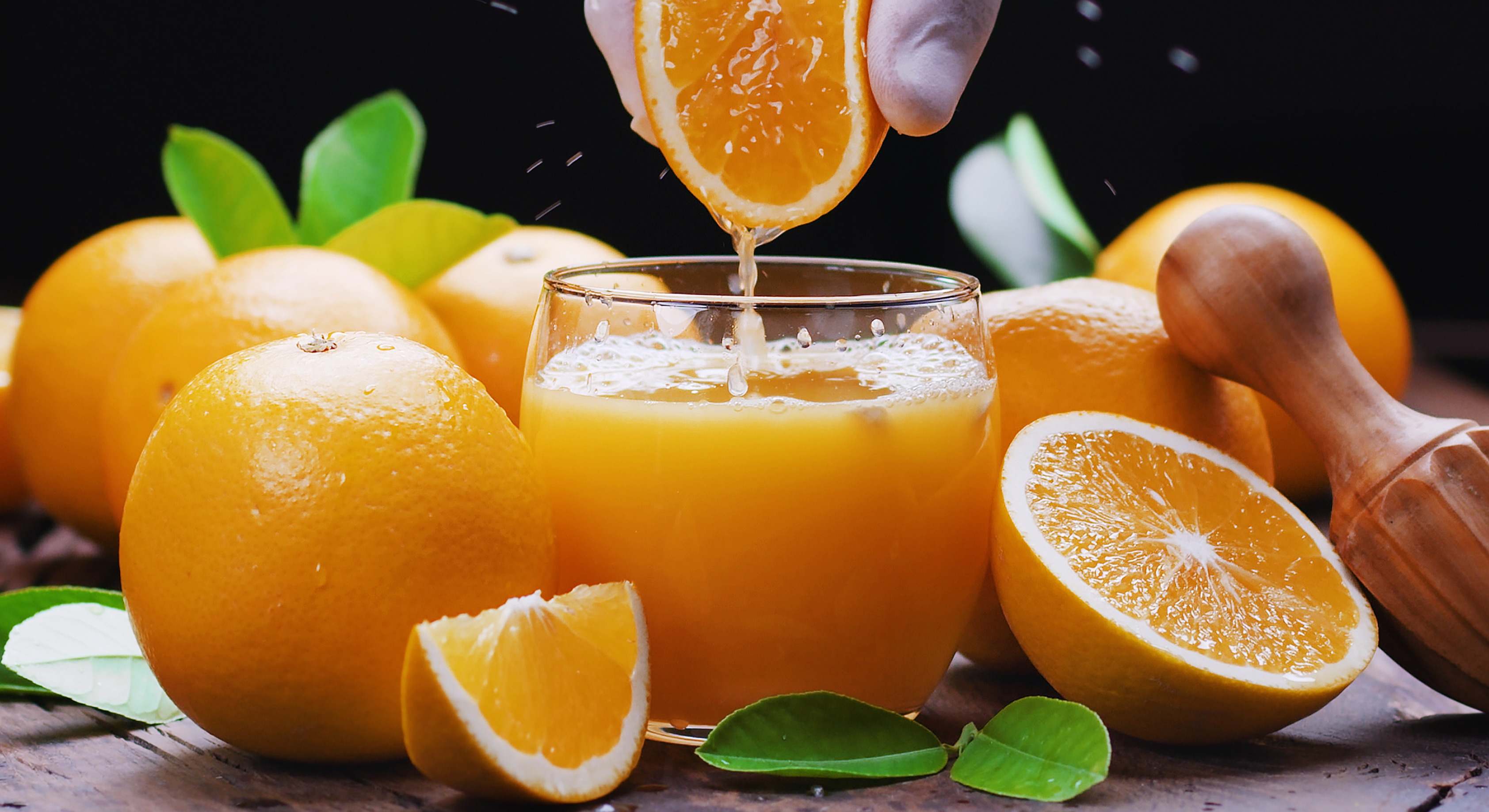 Persona exprimiendo una naranja sobre un vaso de cristal rodeado de más naranjas