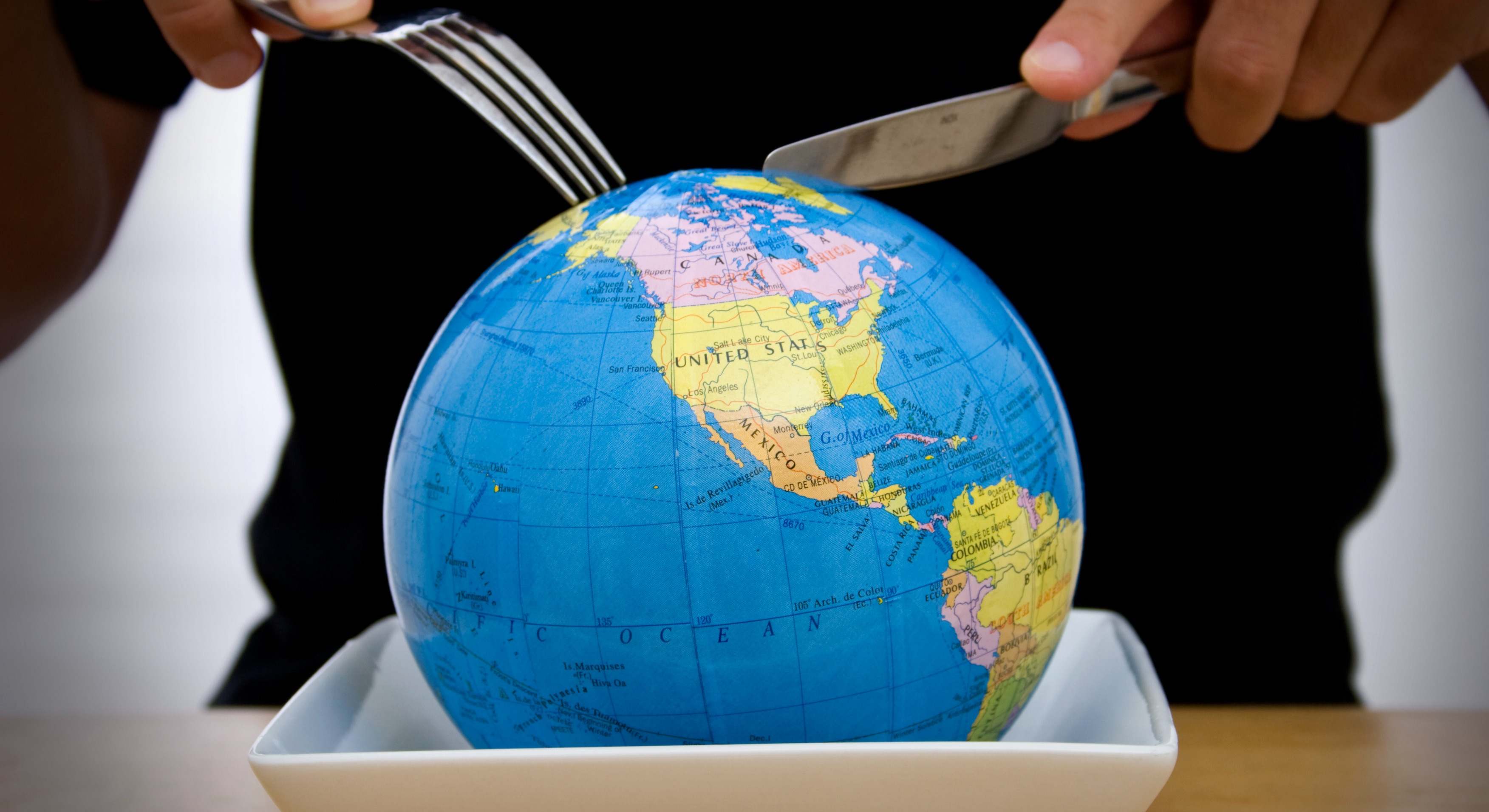 Persona con cuchillo y tenedor cortando un globo terráqueo sobre un plato