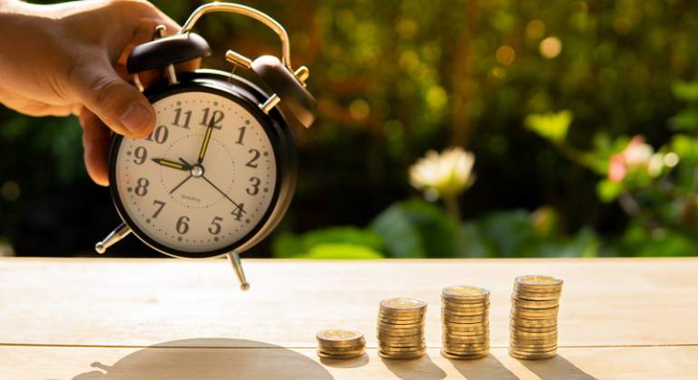 Una mano sujeta un reloj despertador cerca de unas monedas