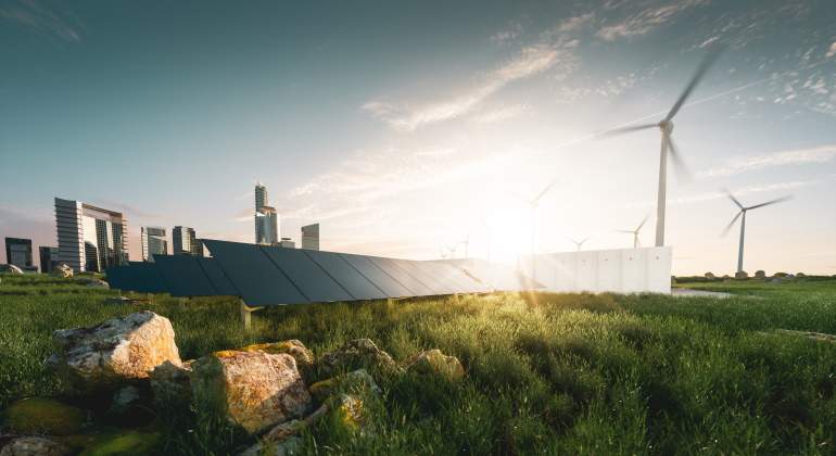 Un campo verde con placas solares y aerogeneradores con la ciudad a lo lejos