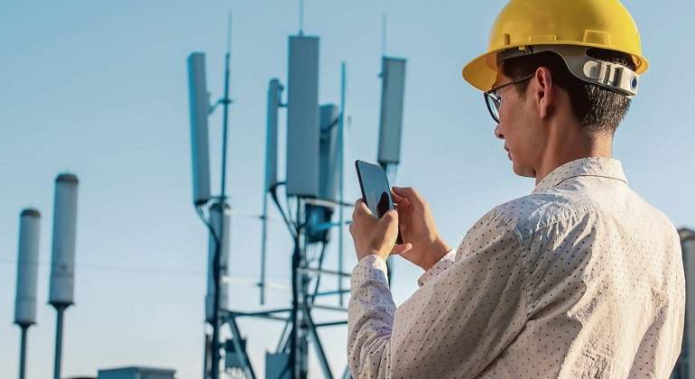 Una persona con casco amarillo sostiene un mvil ante lo que parecen antenas de telefona